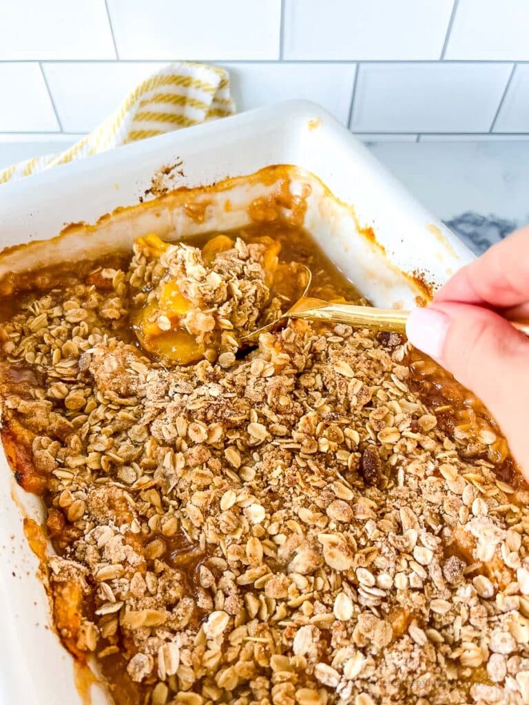 a casserole dish with peach crisp and a hand holding a golden spoon scooping the crisp.