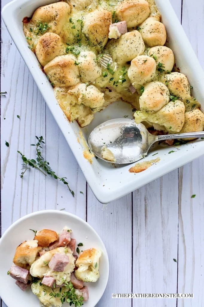 white casserole dish of ham and brie pull apart with a serving spoon and a plate of the ham and cheese casserole