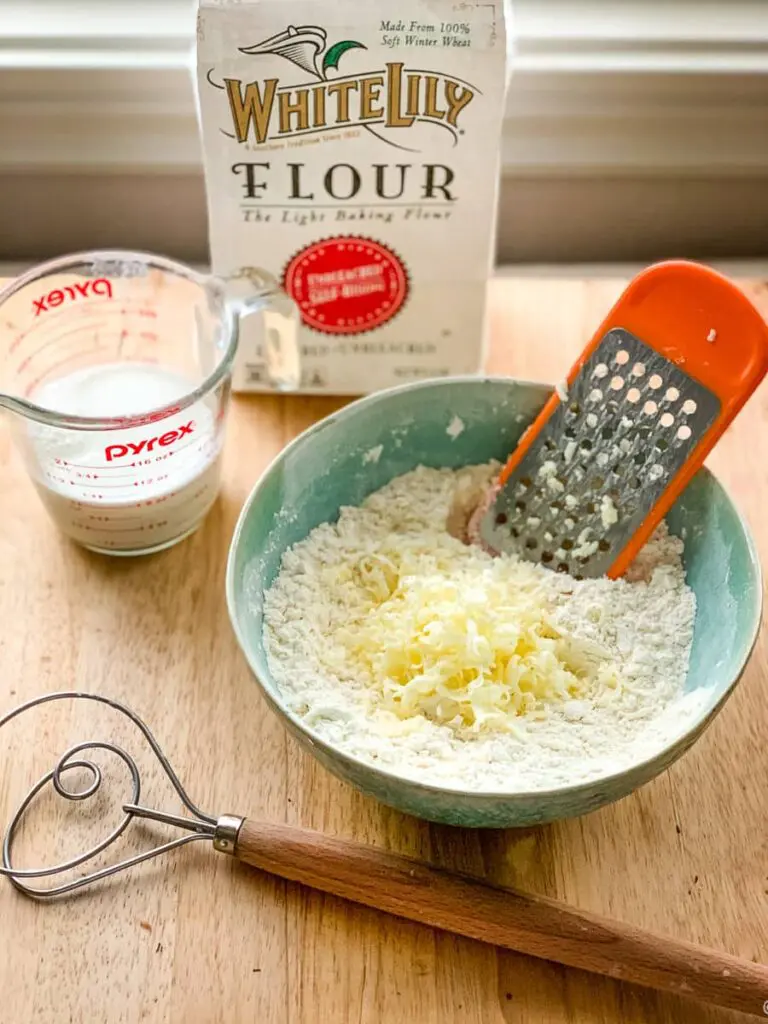 blue bowl of grated butter and self rising flour with buttermilk, a bag of self rising flour, and a dough whisk shown.