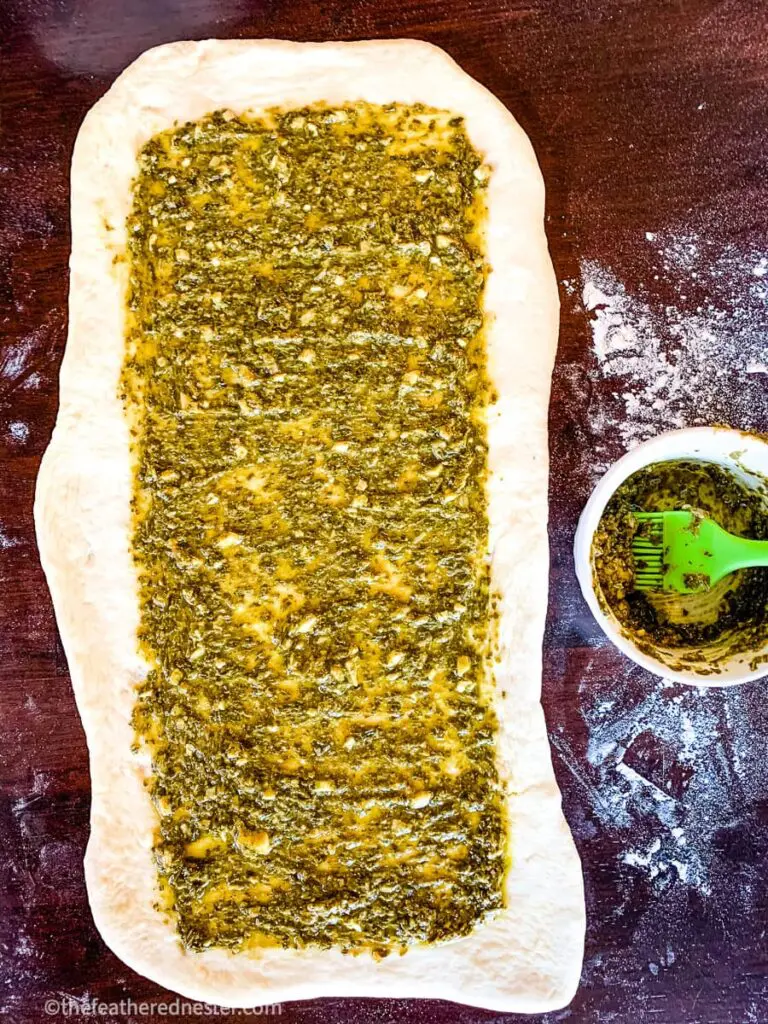Rectangle-shaped piece of dough, covered partially with green sauce, sitting on a lightly floured countertop.