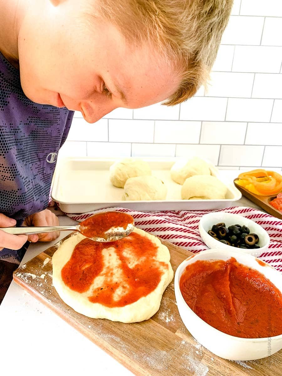 a boy spreading pizza sauce on pizza dough.
