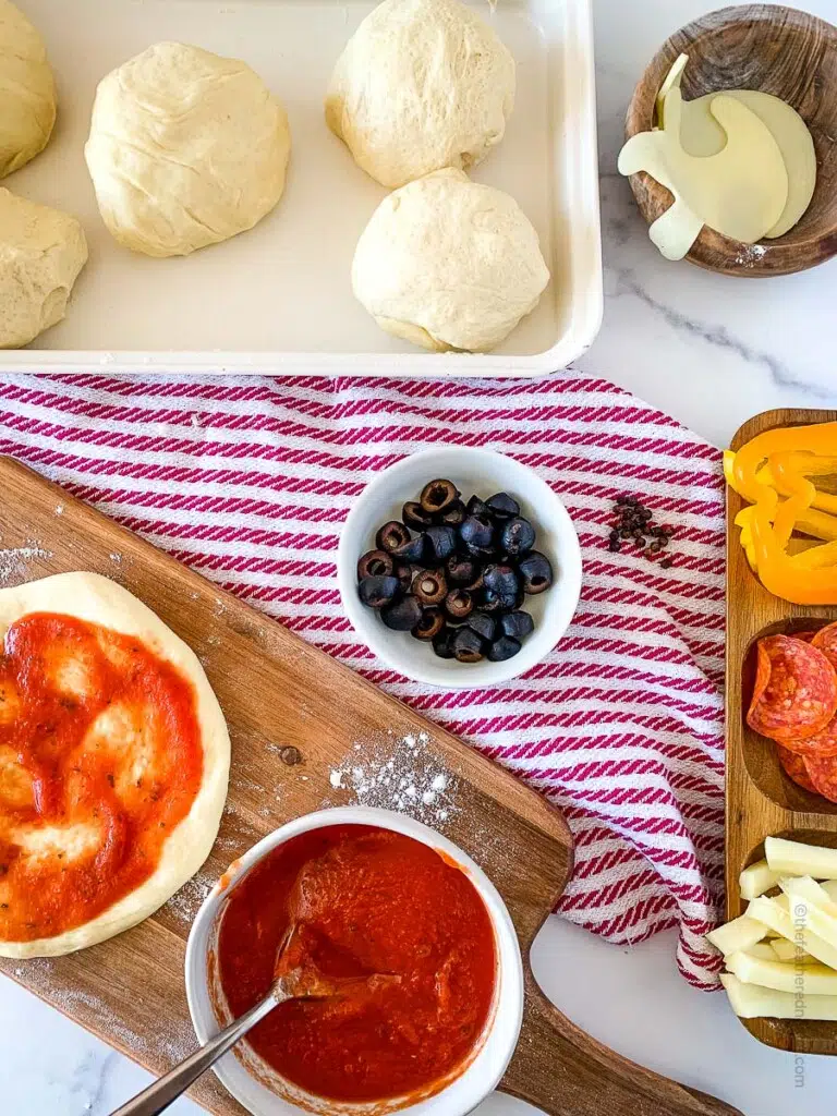 the ingredients to make mini pizzas: balls of pizza dough on a baking sheet, mozzarella ghost cut outs, pizza sauce, and cut vegetables