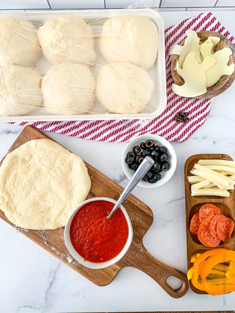 the ingredients to make mini pizzas: balls of pizza dough on a baking sheet, mozzarella ghost cut outs, pizza sauce, and cut vegetables