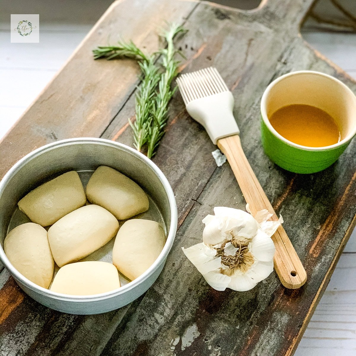 a wooden board with a frozen bread dough rolls in a round baking dish with a pastry brush and green bowl of melted butter and head of garlic.