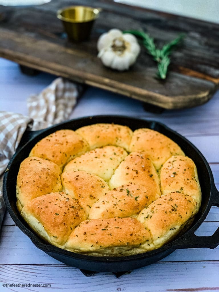 Cast iron skillet with rosemary garlic dinner rolls and a head of garlic, rosemary in the background