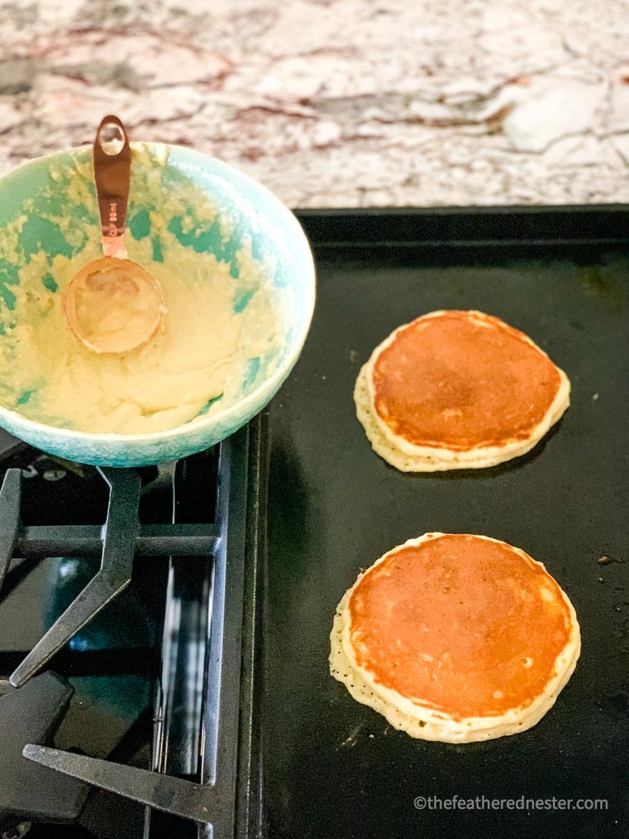Ultimate Bisquick pancakes cooking on a griddle with a bowl of batter.
