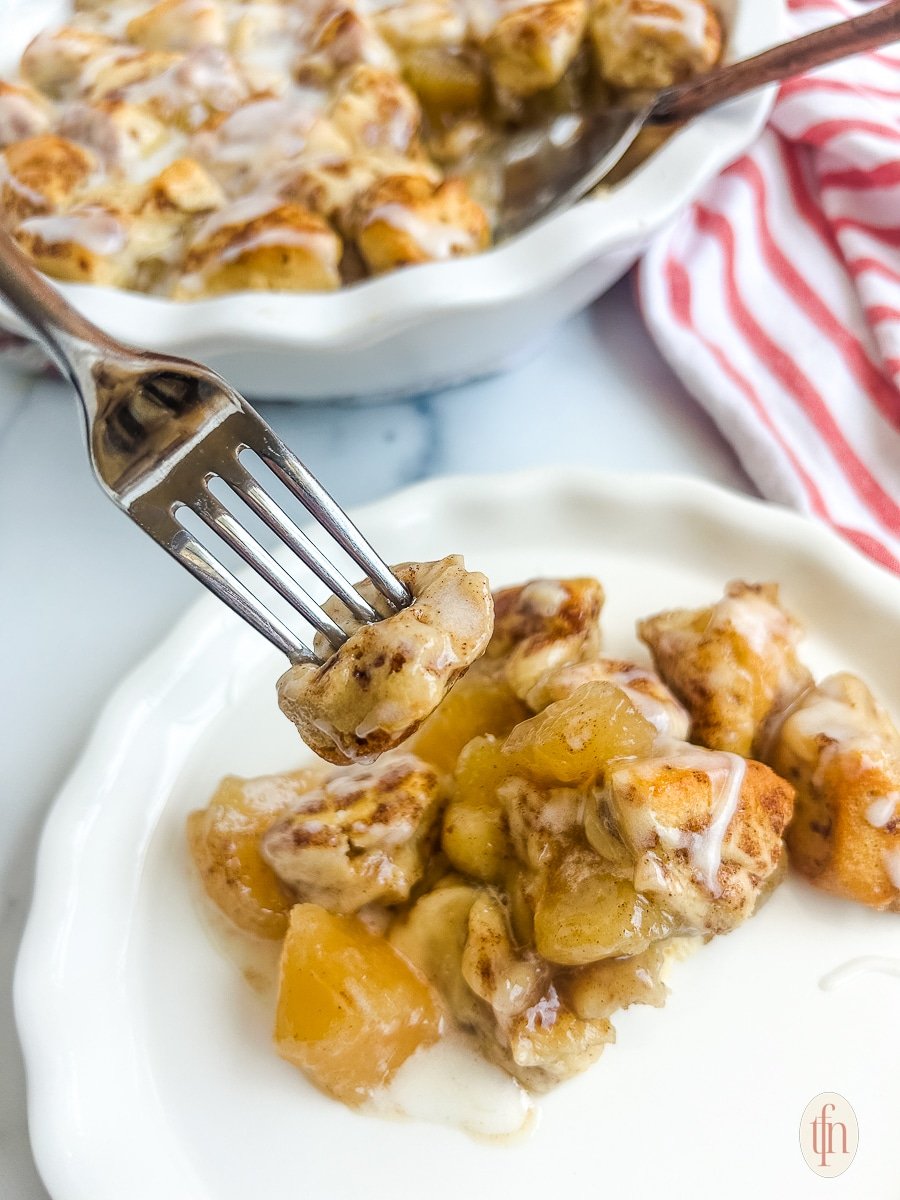 Pieces of apple pie filling and cinnamon roll on a fork held above a white plate. 