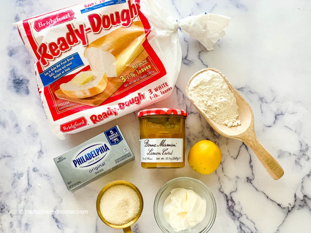 Package of ready-dough frozen bread dough, cream cheese, jar of lemon curd, wooden scoop of flour, gold scoop of sugar, clear bowl of sour cream, and a lemon