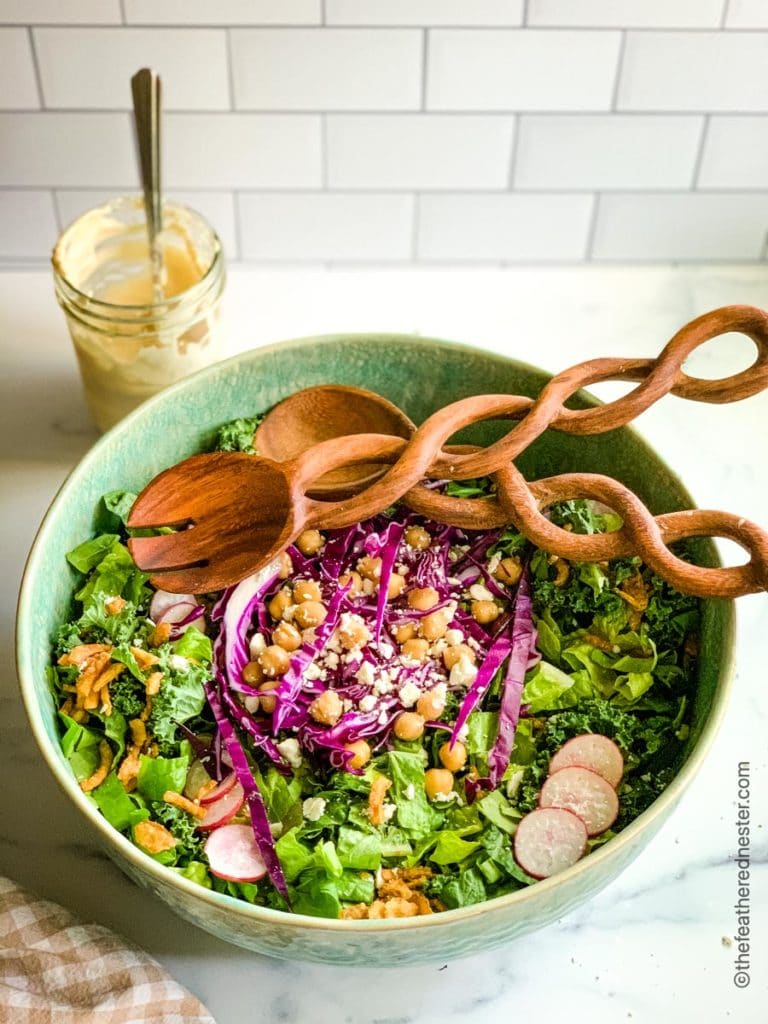 Healthy fresh greens in a green bowl with radishes, chickpeas, feta, and wood tongs. A jar of dressing sits behind the bowl.