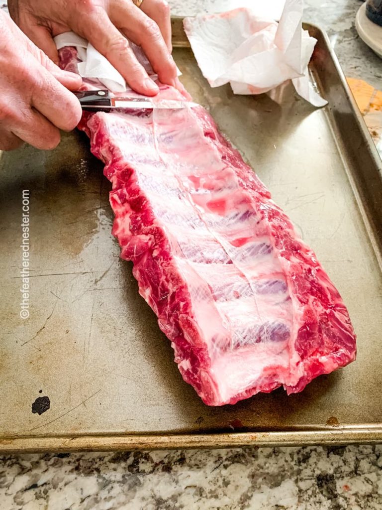 a baking sheet with a rack of baby back ribs bottom side up, a sharp paring knife is being used to separate the membrane from the rib bones