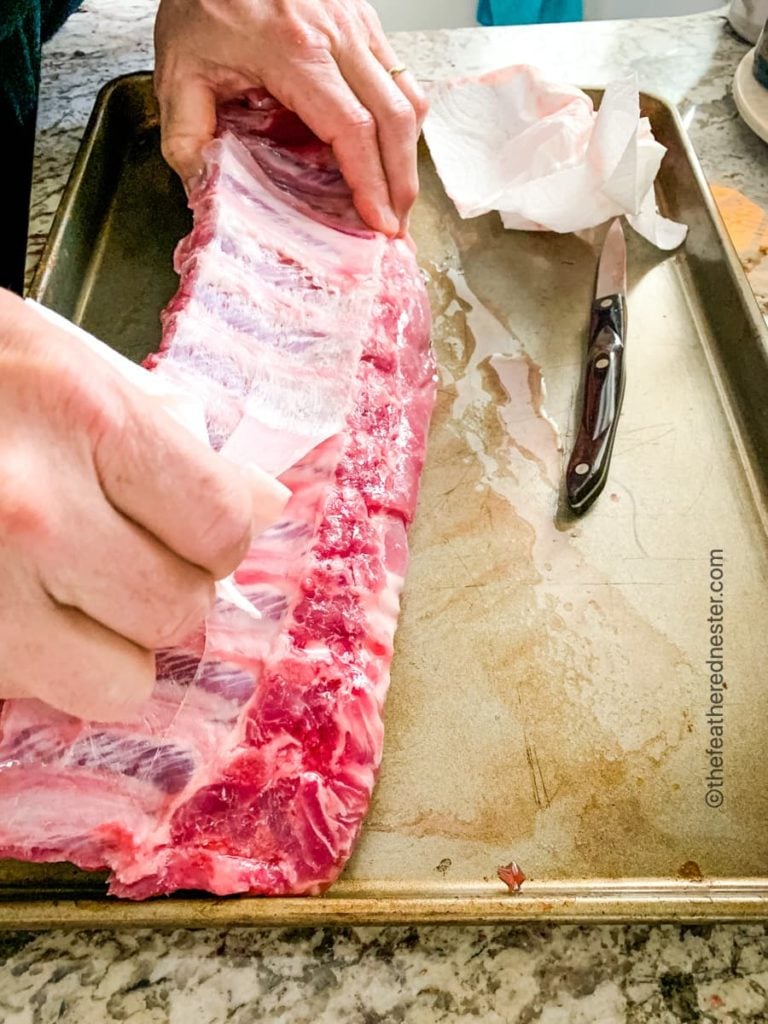 a baking sheet with a rack of baby back ribs, the membranes are being stripped from the rib bones