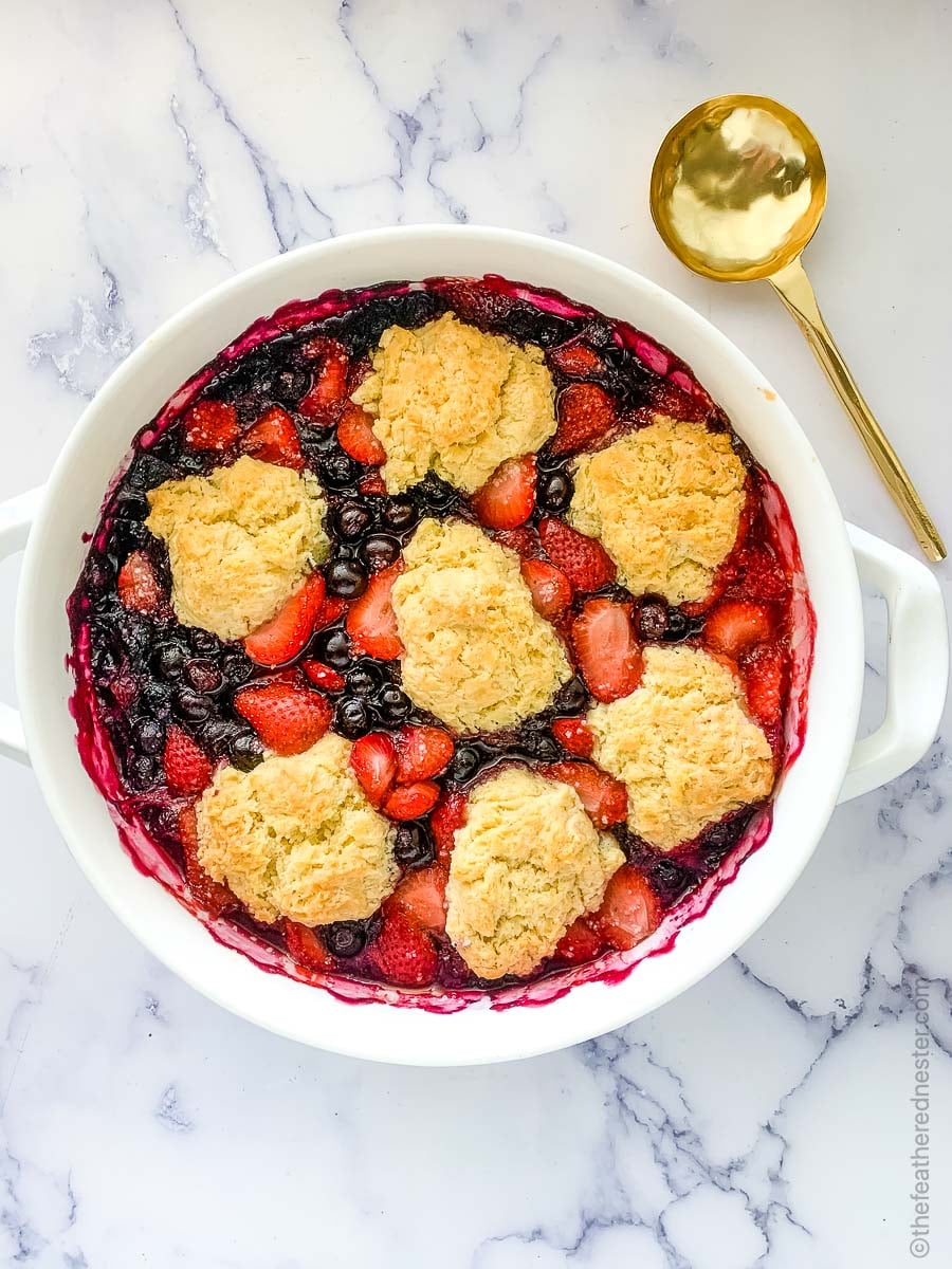 A white casserole dish of baked strawberry blueberry cobbler ready to serve with a gold serving spoon.