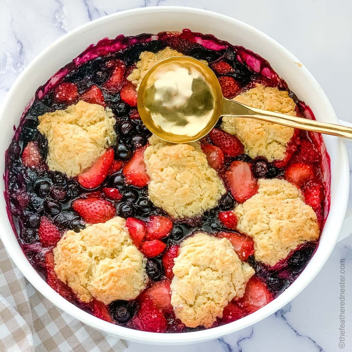 a white casserole dish with strawberry and blueberry cobbler and a gold serving spoon.