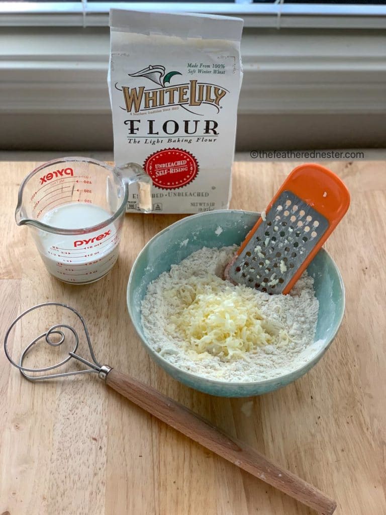 Package of self rising flour next to a blue bowl with grated butter, a measuring cup with buttermilk, and a whisk.