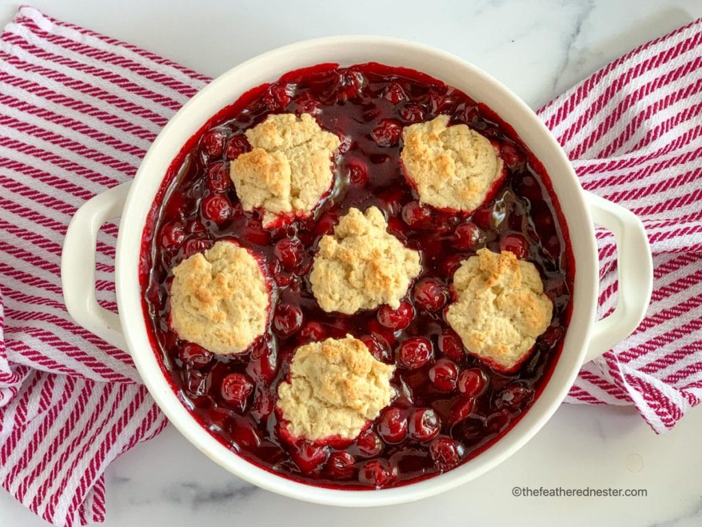 Baked Bisquick cherry cobbler in round white baking dish, sitting on a red and white striped napkin.