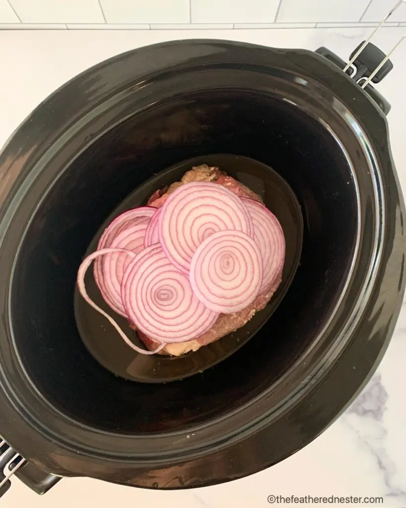 the crock pot roast with ranch dressing is in the slow cooker, covered with sliced onions in beef broth