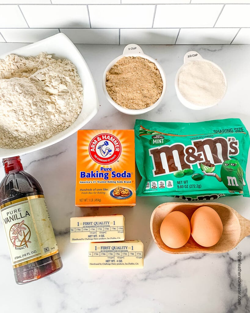 Ingredients on a counter top to make M&M christmas cookies:  mint-flavored candies, eggs, butter baking soda, brown sugar, flour, sugar, vanilla