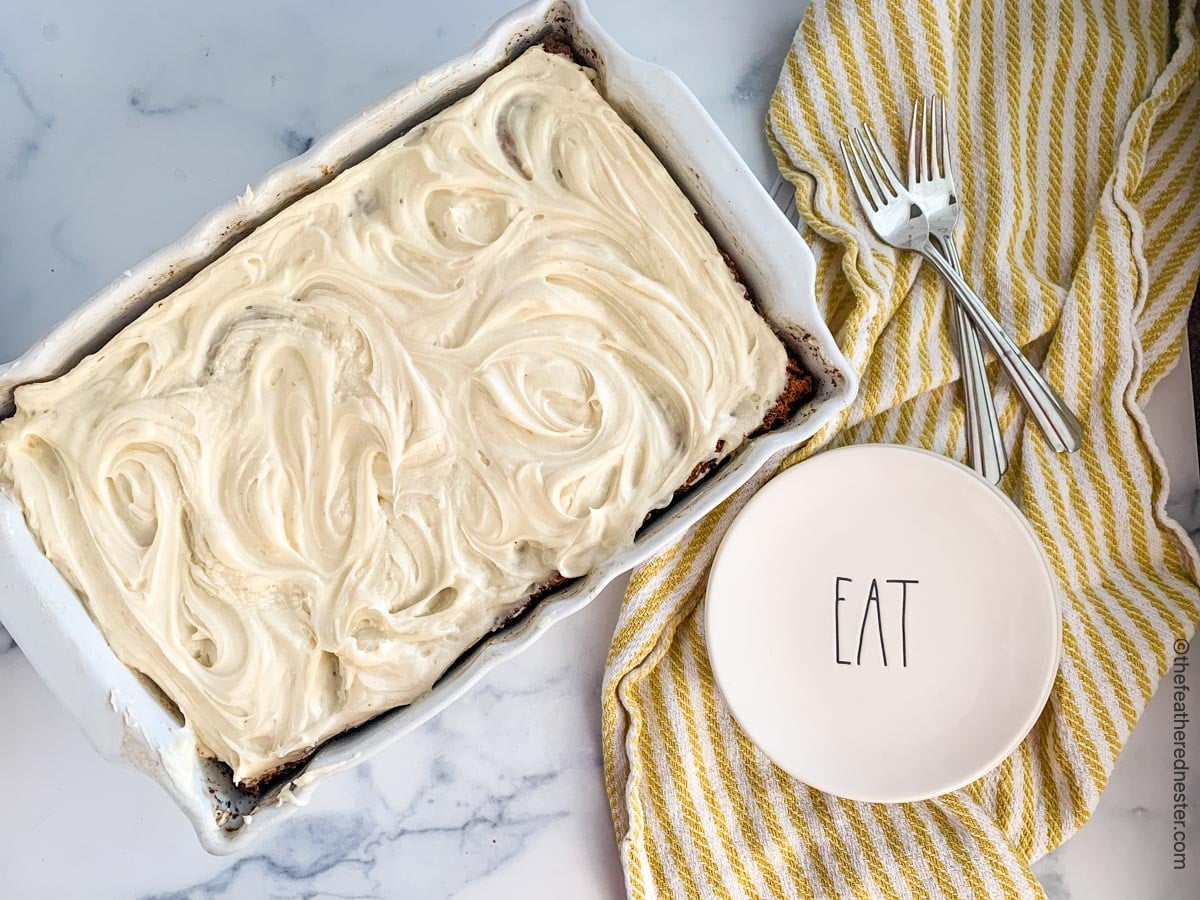 A white pan of sourdough cake with small dessert plates and forks on top of a yellow and white kitchen towel next to it.