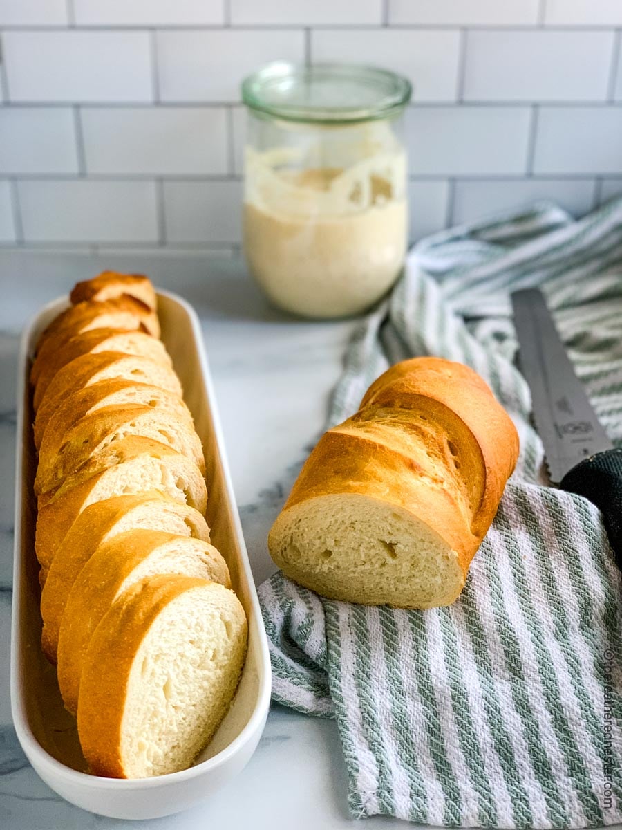 slices of Sourdough French Bread in a white dish with a partially cut loaf on a green and white striped napkin and glass jar of starter in the background.