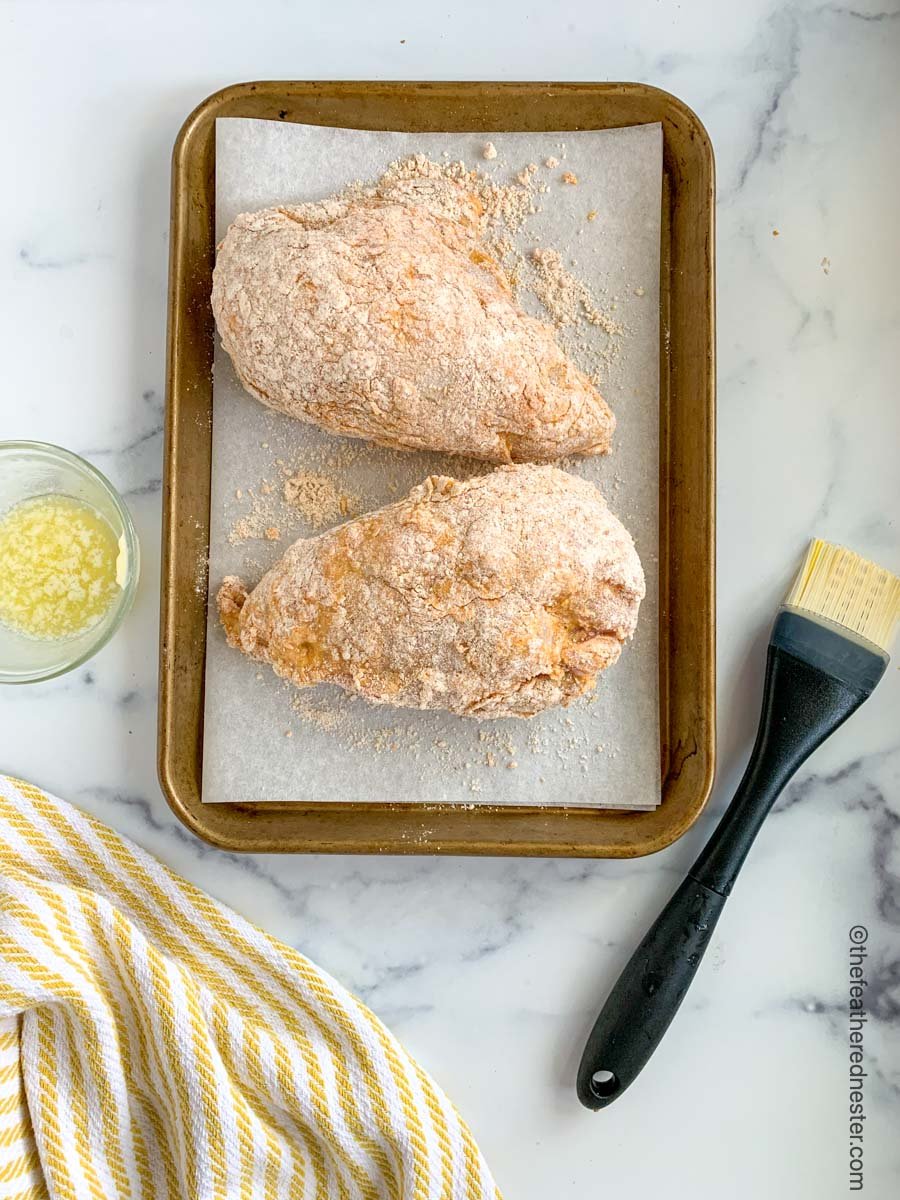 A baking sheet lined with parchment paper and two chicken breasts dipped in bisquick mix with a basting brush and a bowl of melted butter.