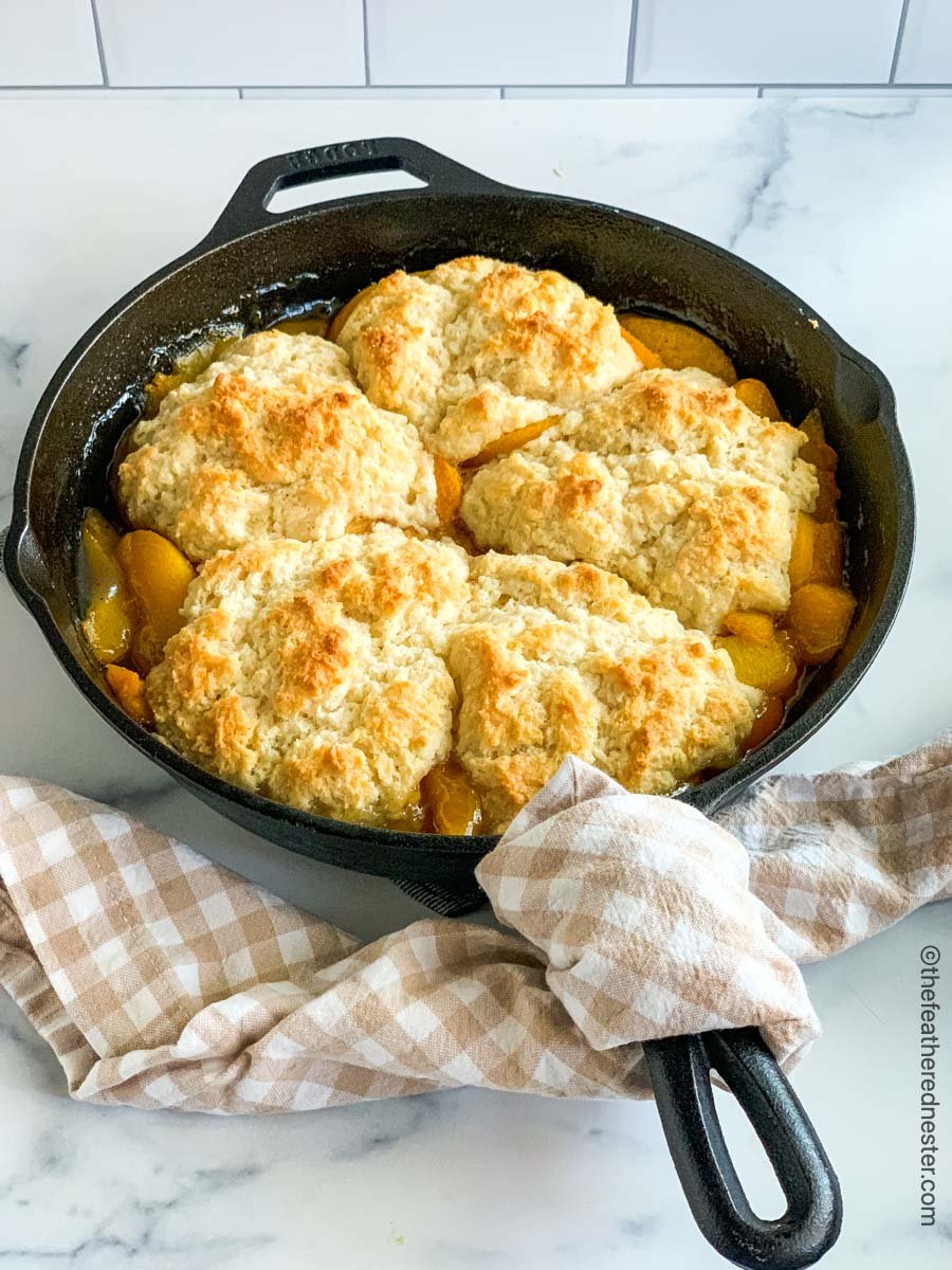 Peach cobbler in a cast iron skillet with a tan and white napkin over the handle.