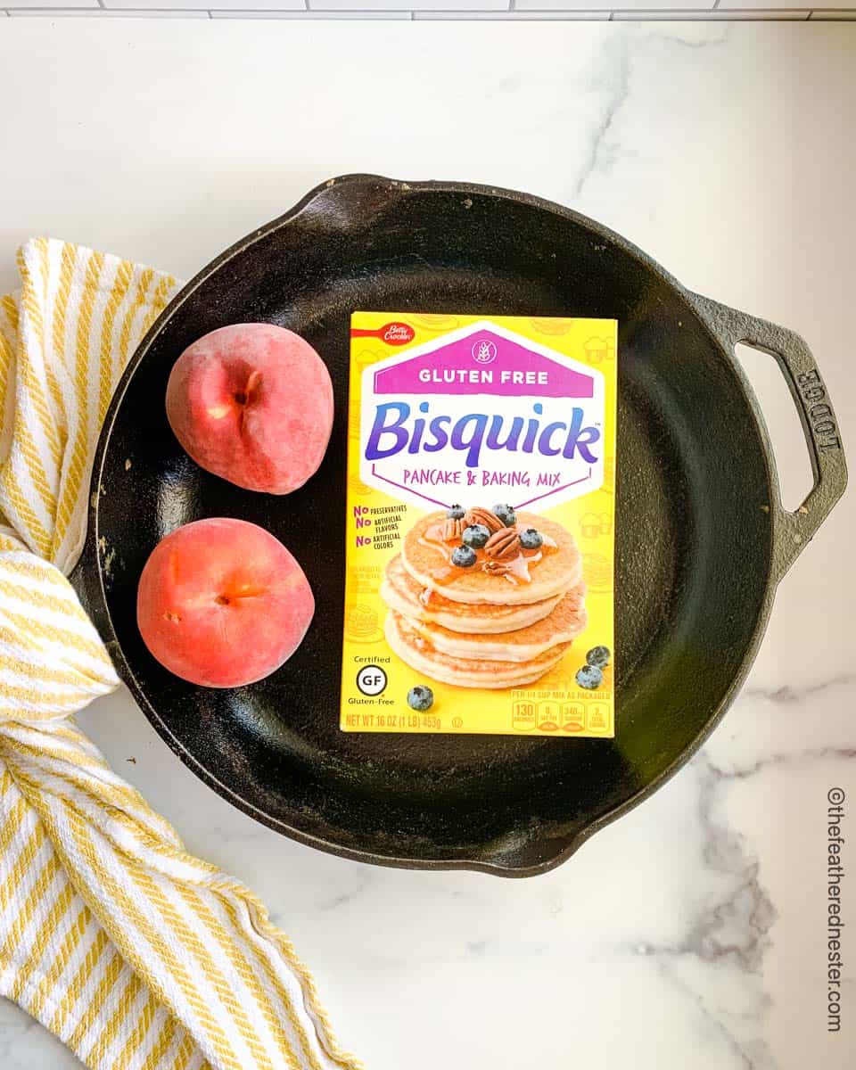 Ingredients for gluten free peach cobbler, a yellow box of gluten free Bisquick and peaches in a cast iron skillet.