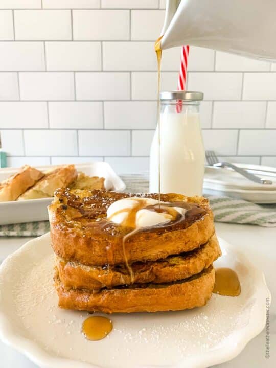 a white plate with a stack of French toast with butter and syrup being poured out of a pitcher over it