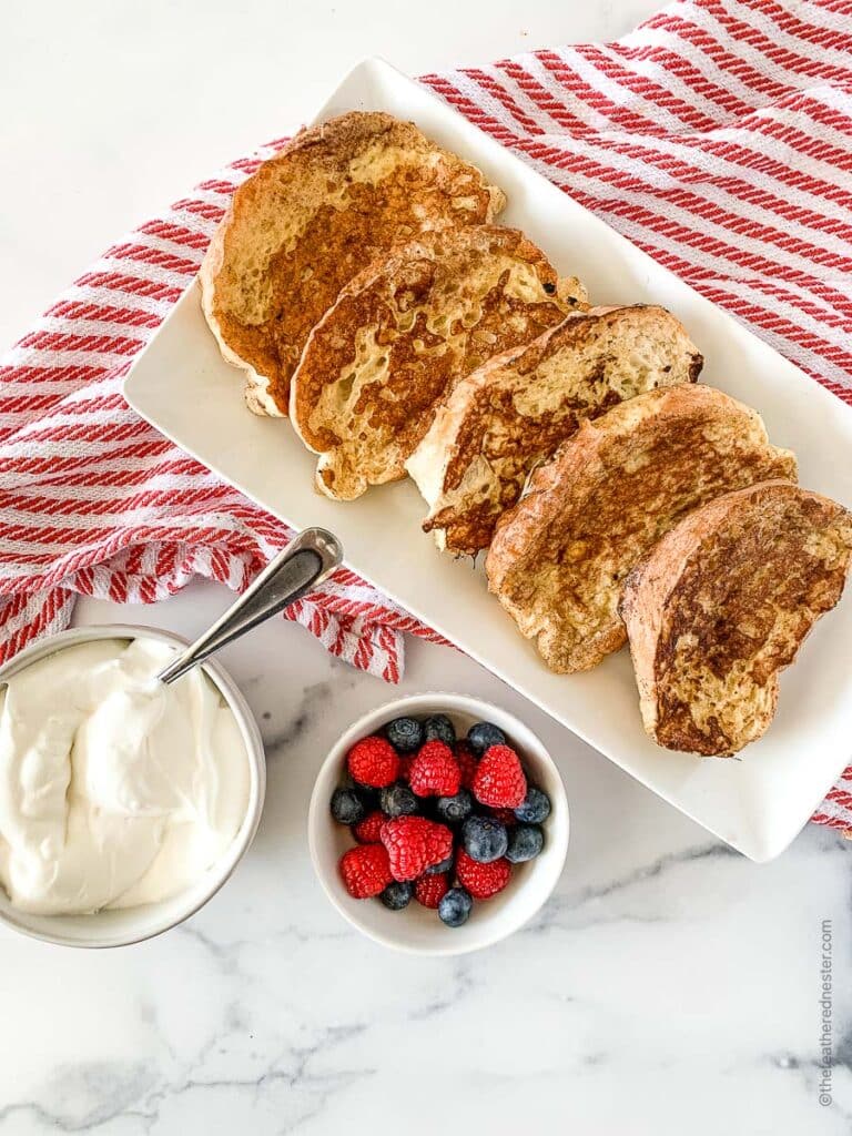 A white platter with slices of sourdough bread French toast, a small bowl of berries and whipped cream over a red and white napkin.