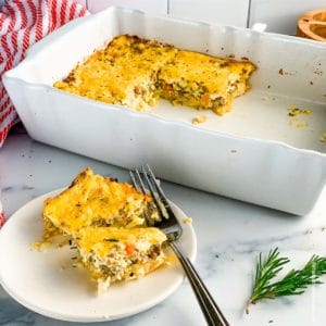 a casserole dish of hash brown casserole with a plate of the egg casserole ready to serve and a striped red towel in the background.