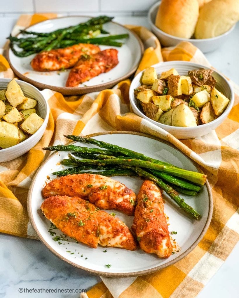 two plates with baked chicken tenders with asparagus, and two plates of baked potatoes.