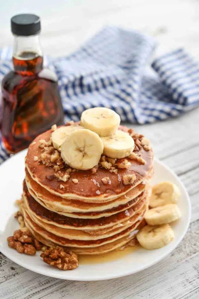 a stack of banana pancakes with nuts and maple syrup in the background