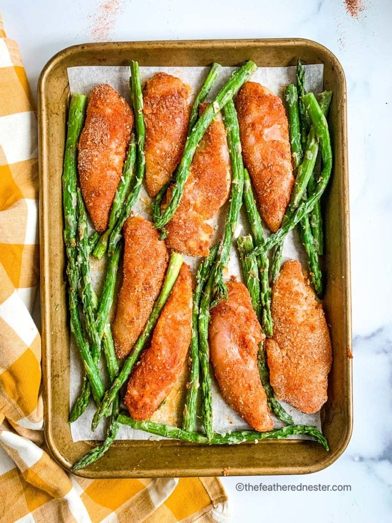A sheet pan of chicken tenderloins dipped in seasonings with asparagus spears