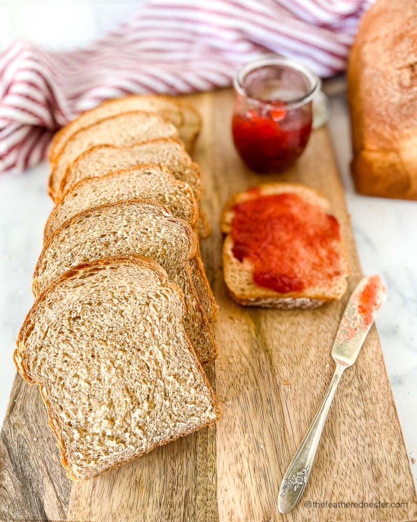 a wooden board with a loaf of sliced wheat bread with a slice of bread spread with strawberry jam and a jam jar