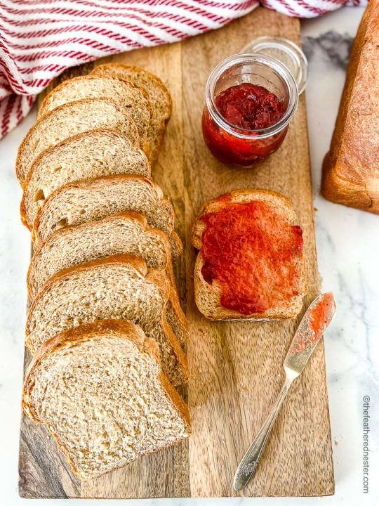 a wooden board with a loaf of sliced wheat bread with a slice of bread spread with strawberry jam and a jam jar