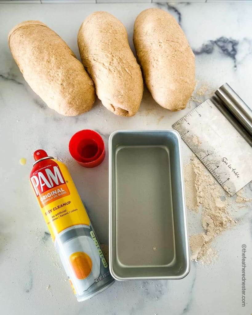 a bread pan, a can of cooking spray, and three loaves of dough rolled up