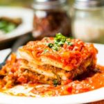 a slice of lasagna with a sprig of basil on top of it on a white place