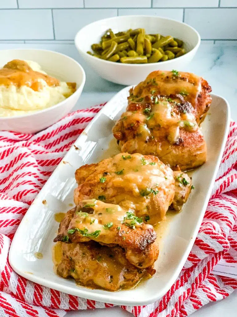 a white platter of chicken thighs with a bowl of green beans and mashed potatoes in the background