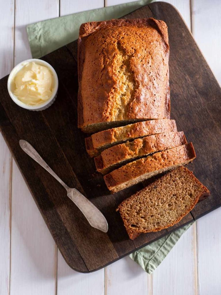 a sliced loaf of sourdough banana bread on a wooden cutting board with a bowl of butter and a bread knife on the left
