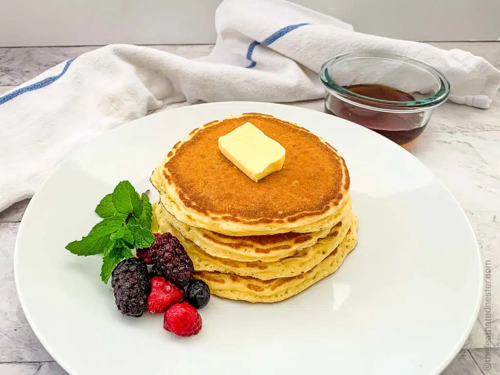 a white plate of pancake garnished with berries and butter topped with a container of syrup in the background