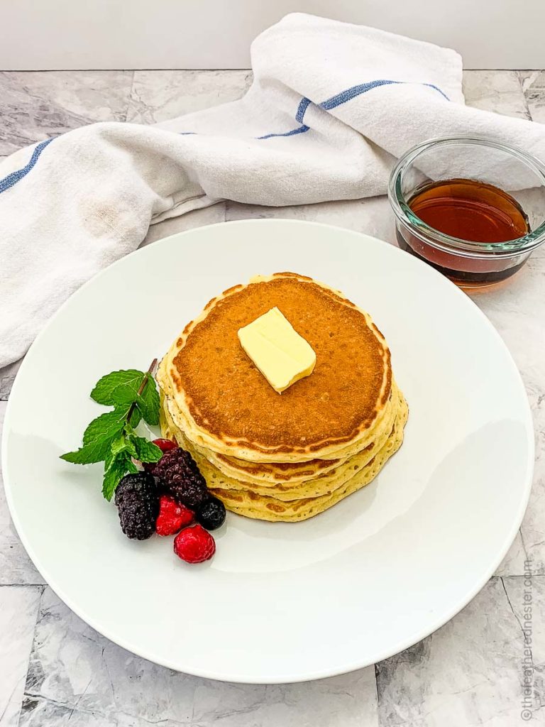 overhead view of a stack of pancakes with berry garnish and butter on a white plate with a container of pancake syrup in the background