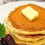 a white plate of pancake garnished with berries and butter topped with a container of syrup in the background