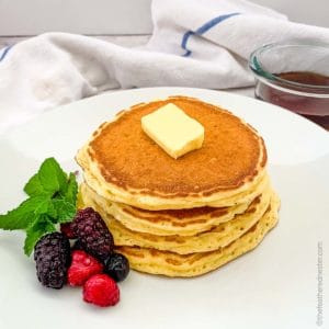 a white plate of pancake garnished with berries and butter topped with a container of syrup in the background.