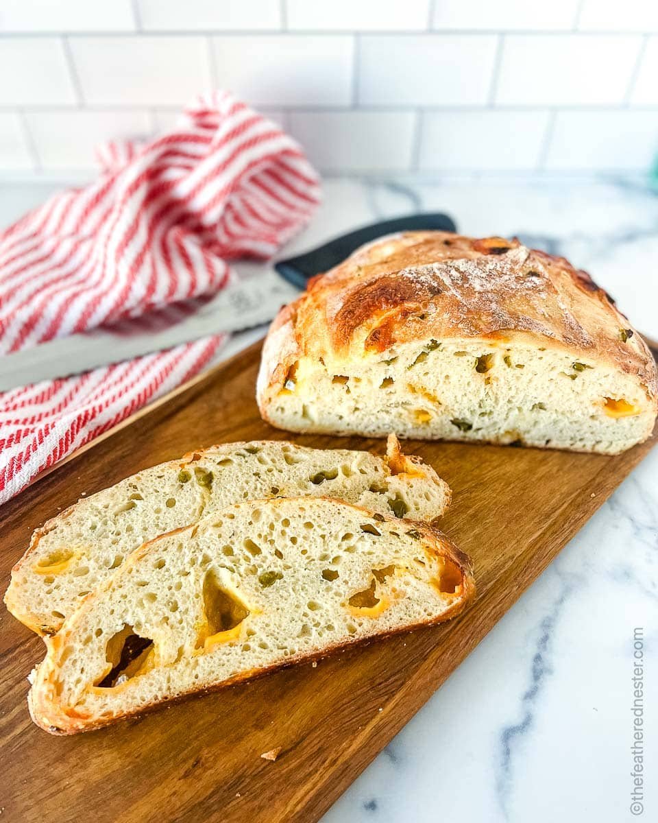 slices of jalapeno cheddar sourdough on a wooden board with the loaf in the background and a red and white towel.