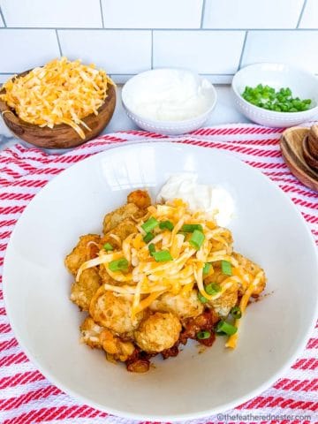 a white plate of chili tater tot casserole topped with shredded cheese and sliced green onions on a red and white napkin with bowls of grated cheese sour cream and sliced green onions in the background