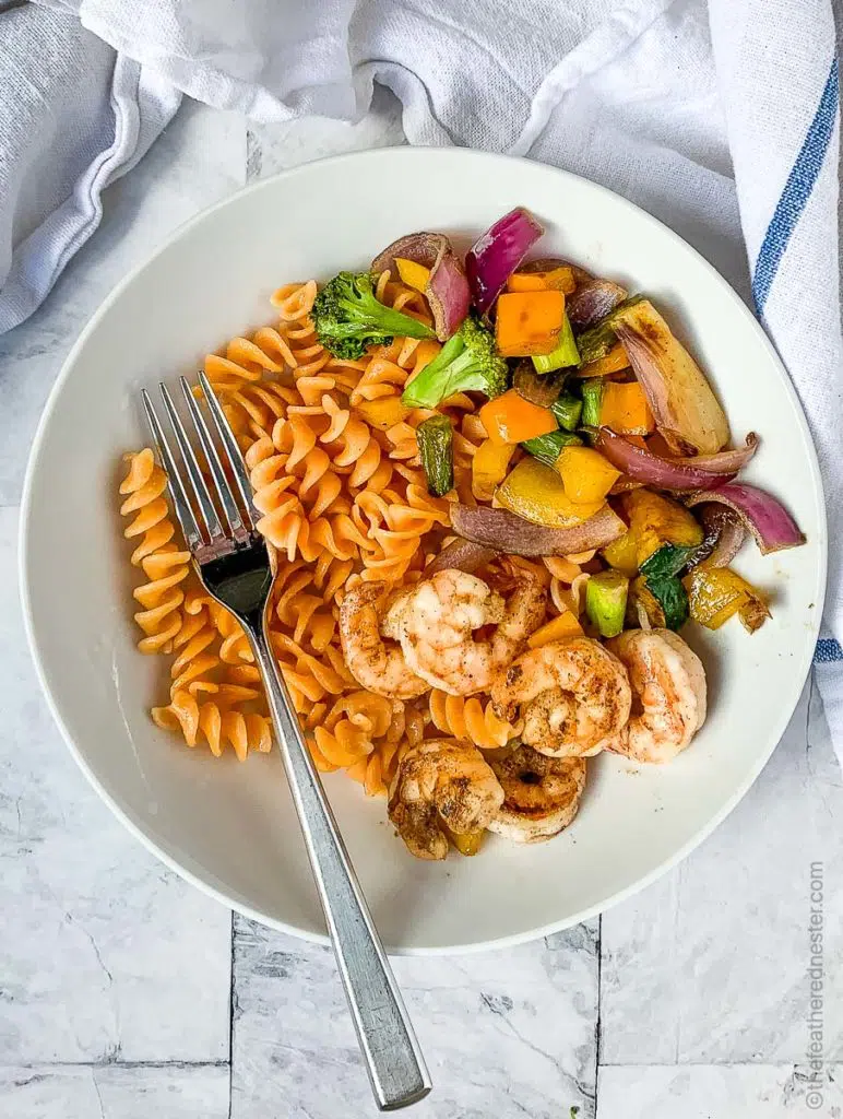 a white plate of veggies, rotini pasta and shrimp with cooked on a flat top grill.