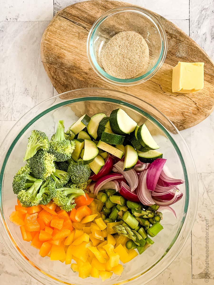 a chopped vegetable medley in a clear glass bowl with a wooden cutting board with a bowl of spices and a pat of butter