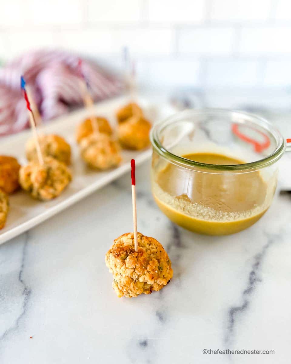 appetizer toothpick stuck in a meatball made from palmetto cheese balls with a dipping sauce in the background and a tray of more appetizer balls.