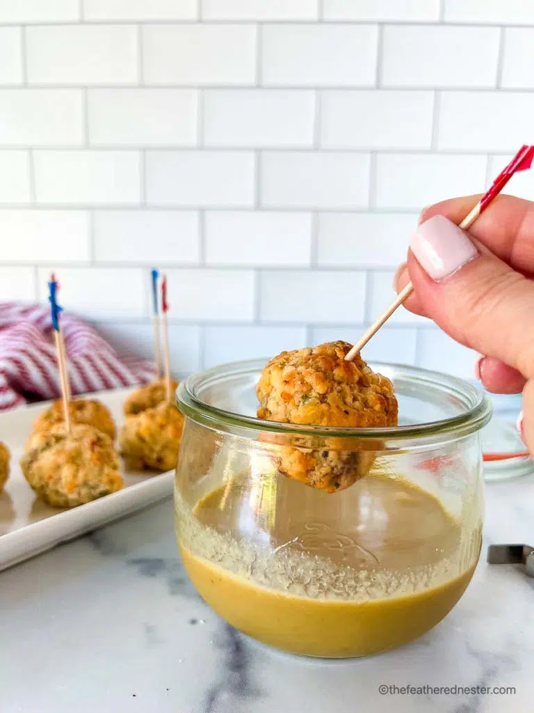 dipping a sausage meat ball being dipped into spicy mustard sauce in a clear glass deck jar with a tray of turkey balls with toothpicks in the background