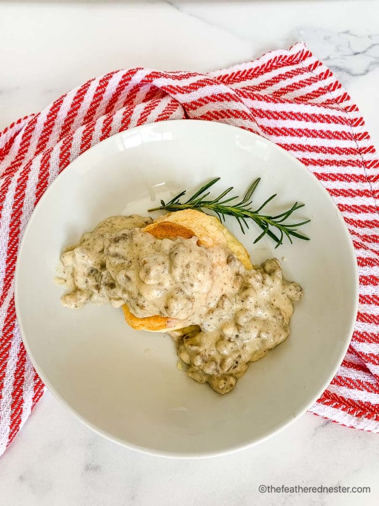 a white plate with turkey gravy and biscuits and a spring of rosemary ready to serve over a red and white napkin