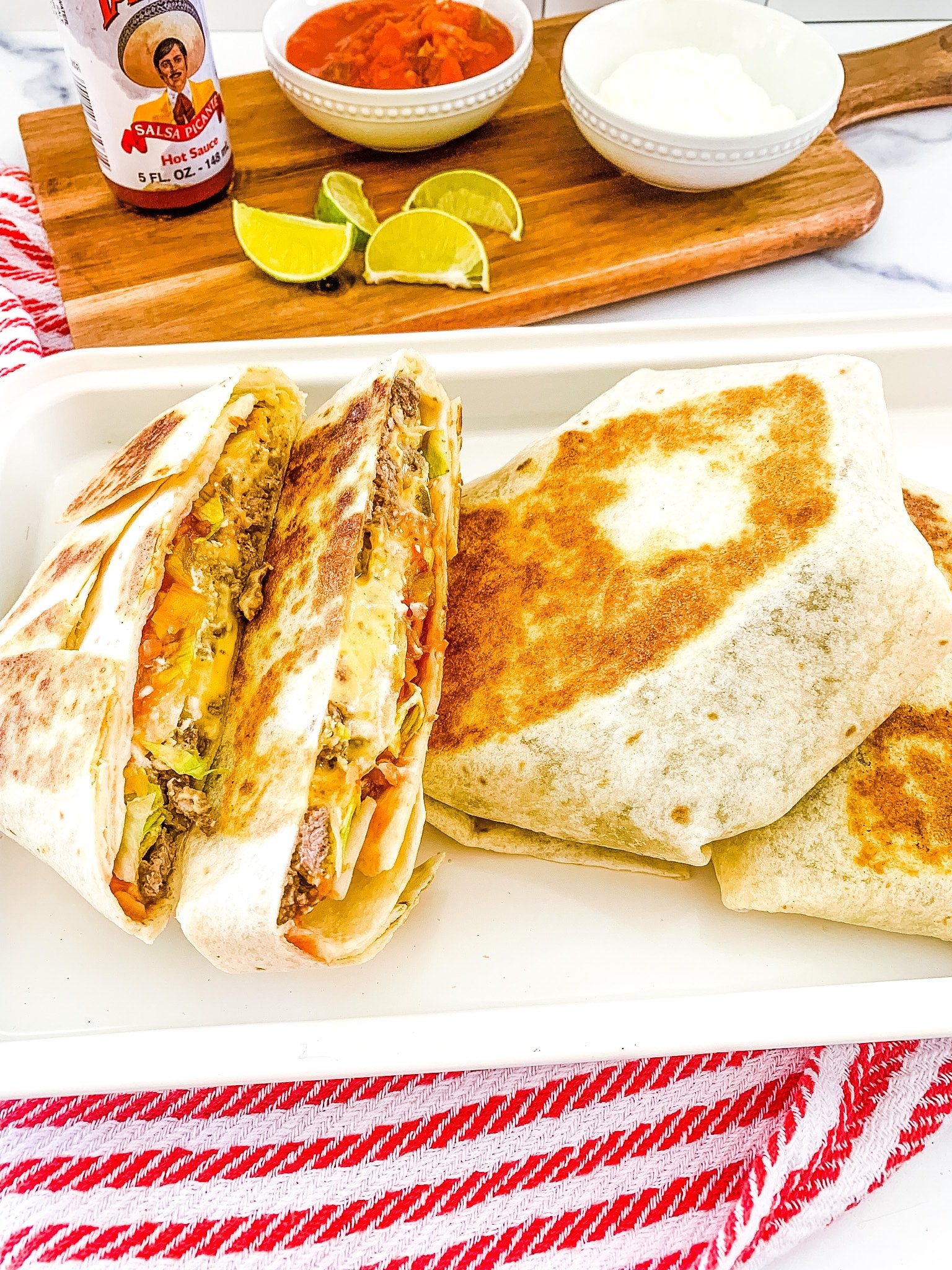 a white platter of Blackstone Crunchwrap tortillas cut in half on top of a red and white napkin, with a bowl of salsa and slices of limes in the background.